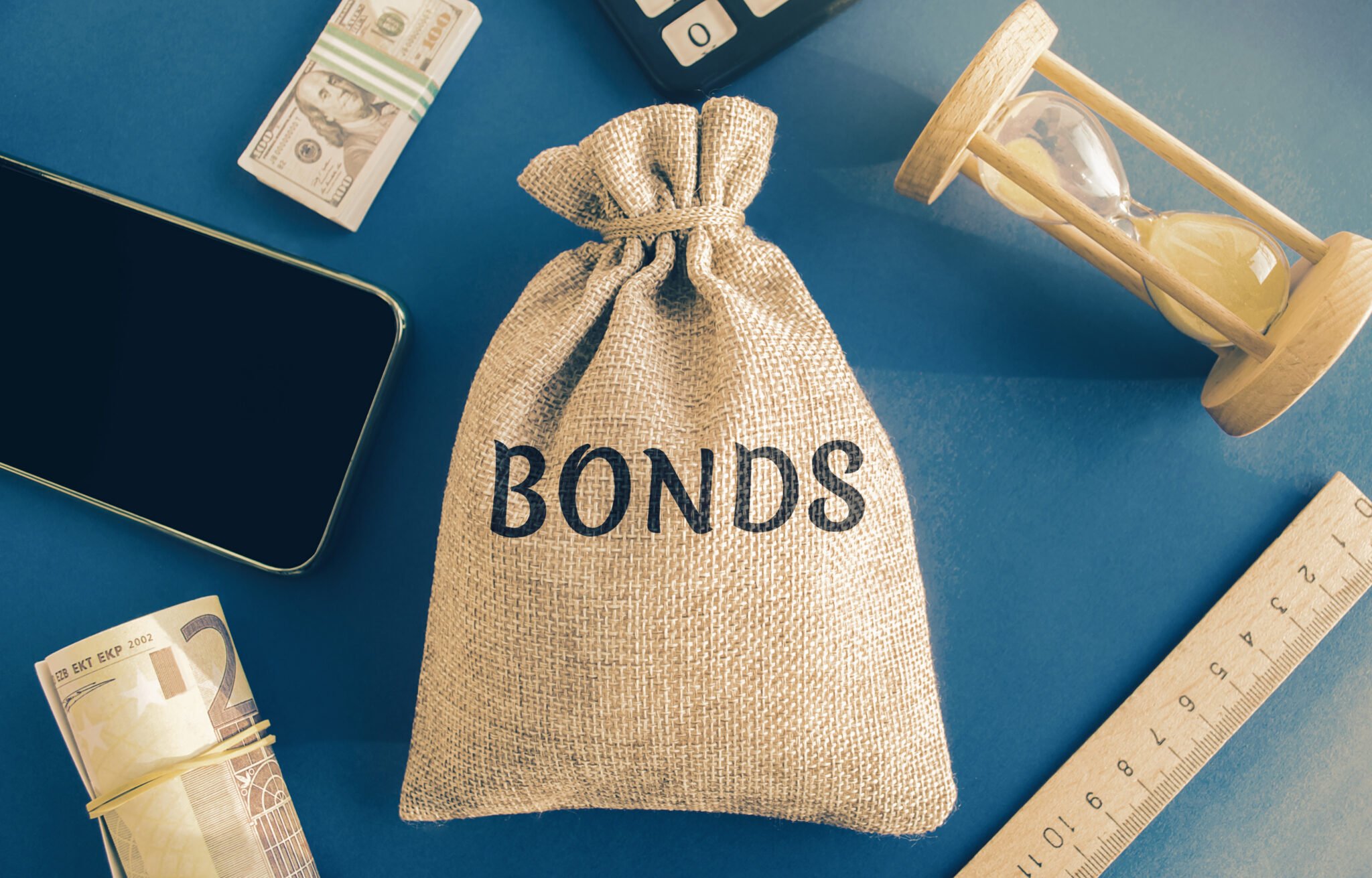 Money bag with the word Bonds. A bond is a security that indicates that the investor has provided a loan to the issuer. Equivalent loan. Unsecured and secured bonds. A burlap bag labeled "Bonds" sits on a table with cash, a phone, an hourglass, and a ruler—perhaps waiting for California's Encinitas Bail Bonds to lend their expertise.