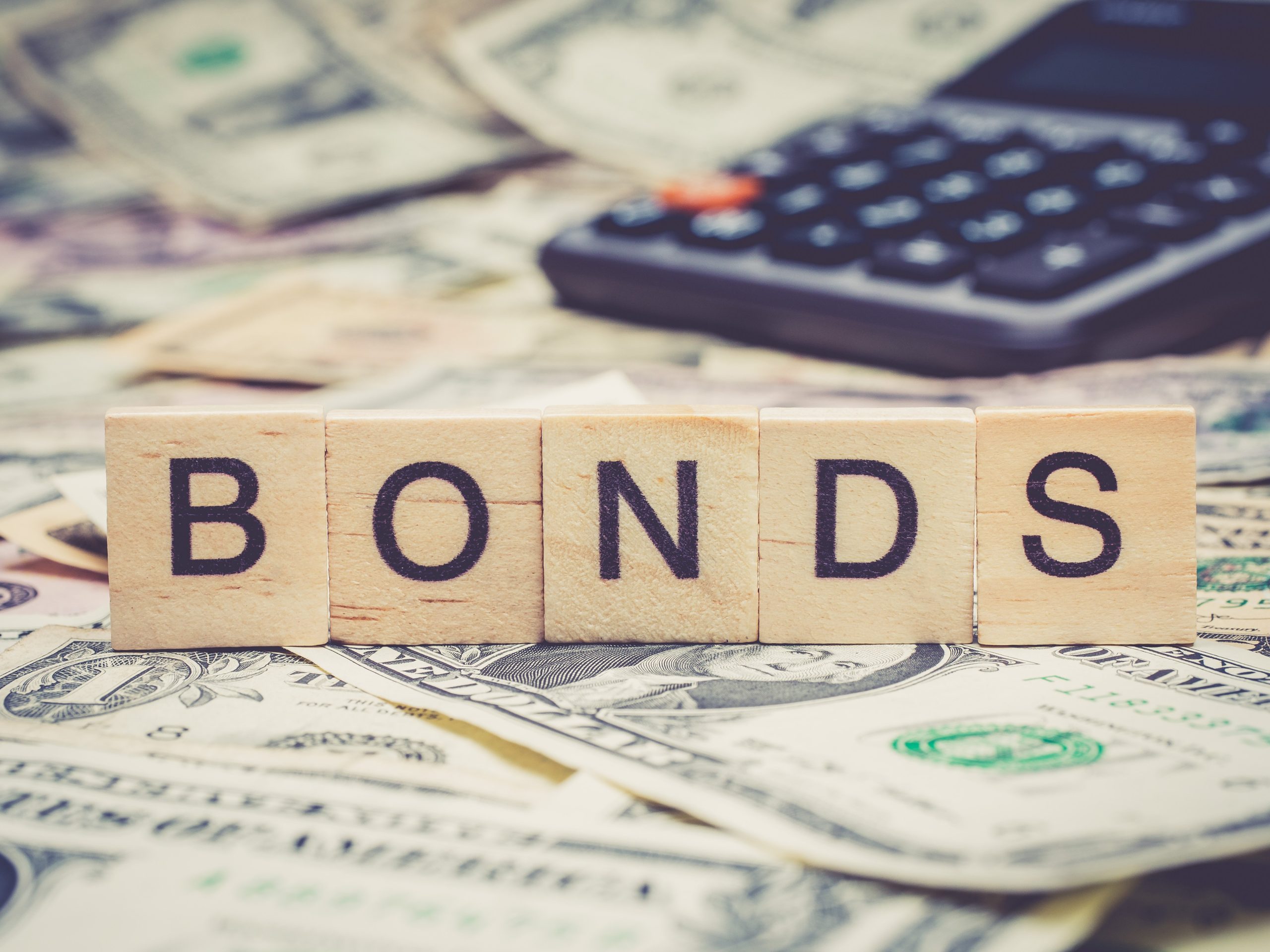 bonds Scrabble tiles spelling "BONDS" lie on a pile of money, with a calculator in the background, subtly hinting at the financial world where terms like Encinitas Bail Bonds play a role.