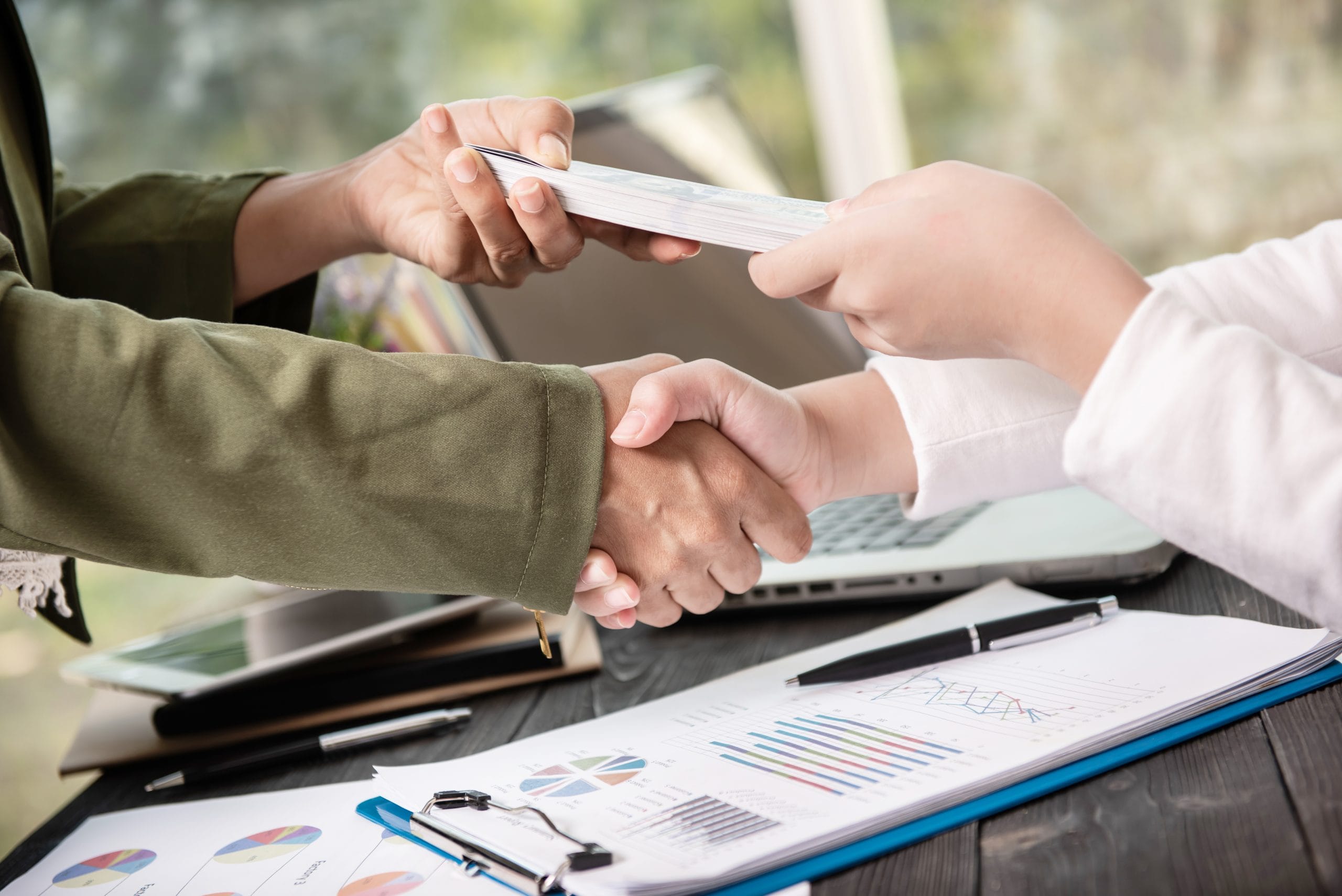 Business people shaking hands, finishing up meeting. businessman Two people shaking hands while exchanging money, with charts and a laptop on a desk in the background.