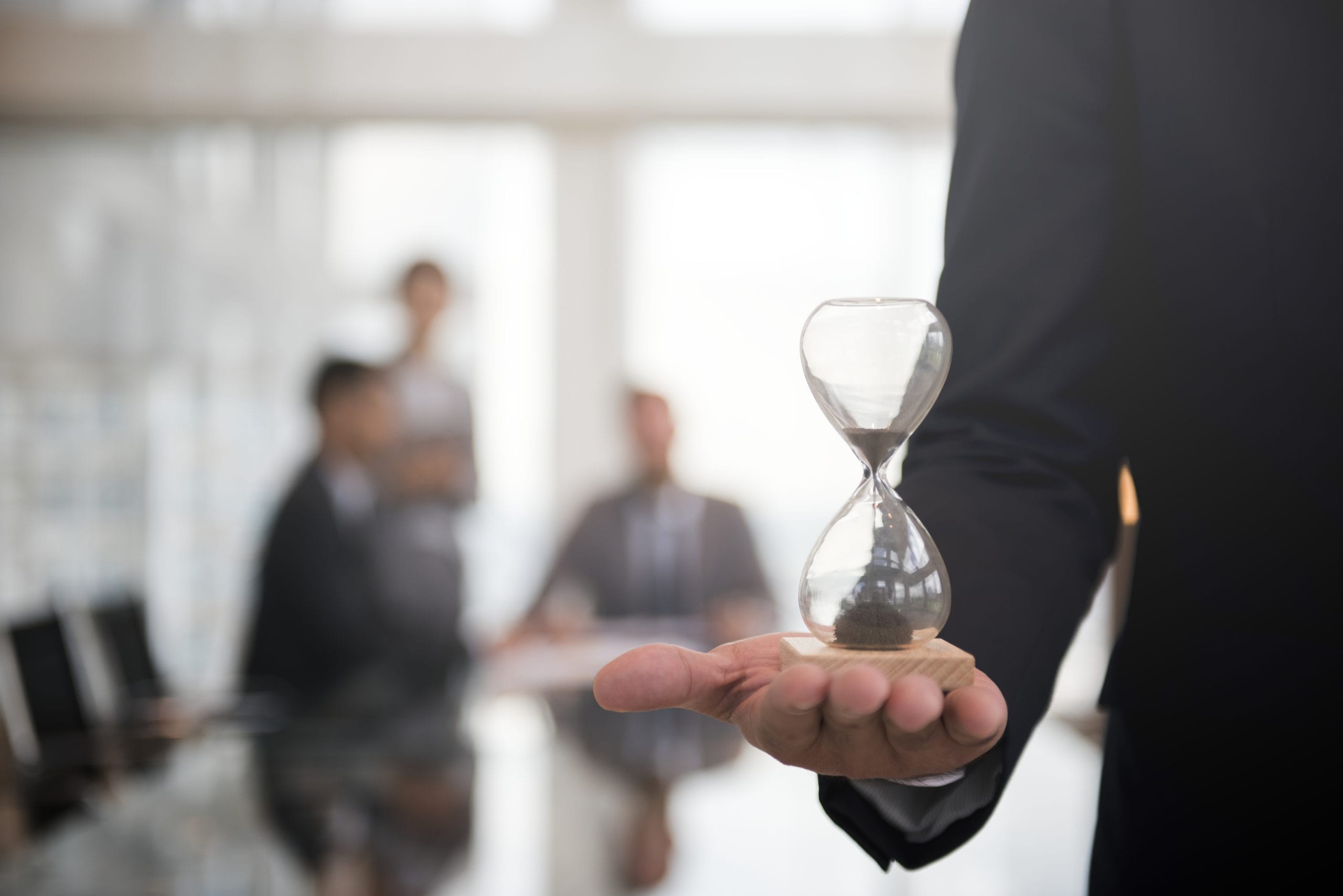 Businessman holding an hour glass, signifies the importance of being on time A person in a suit holds an hourglass, with blurred colleagues in a meeting room in the background.