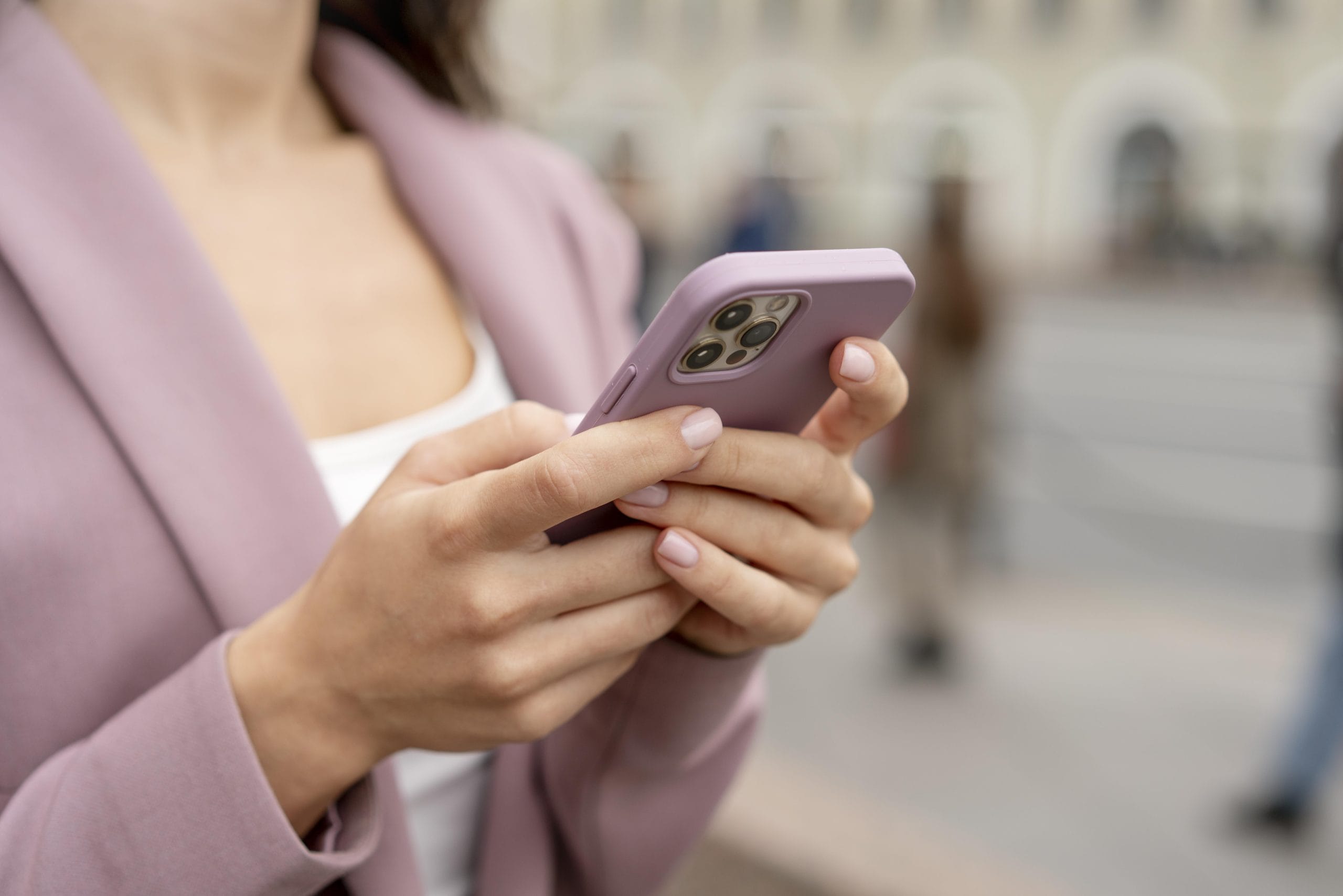 close-up-hands-holding-smartphone Woman in a pink blazer holding and using a smartphone outdoors, background is out of focus.