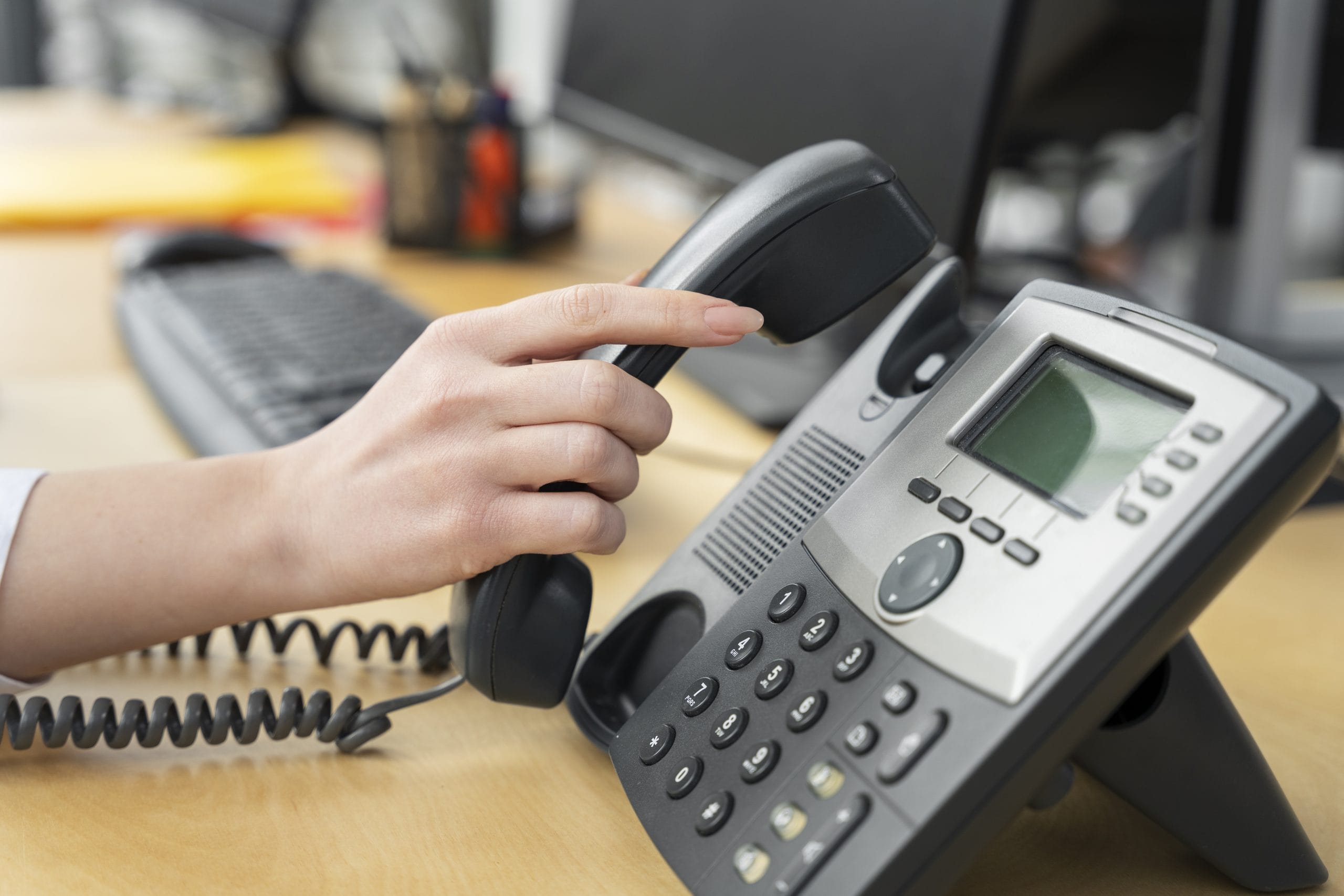 close-up-person-working-call-center A person’s hand picking up the handset of an office desk phone next to a computer keyboard.