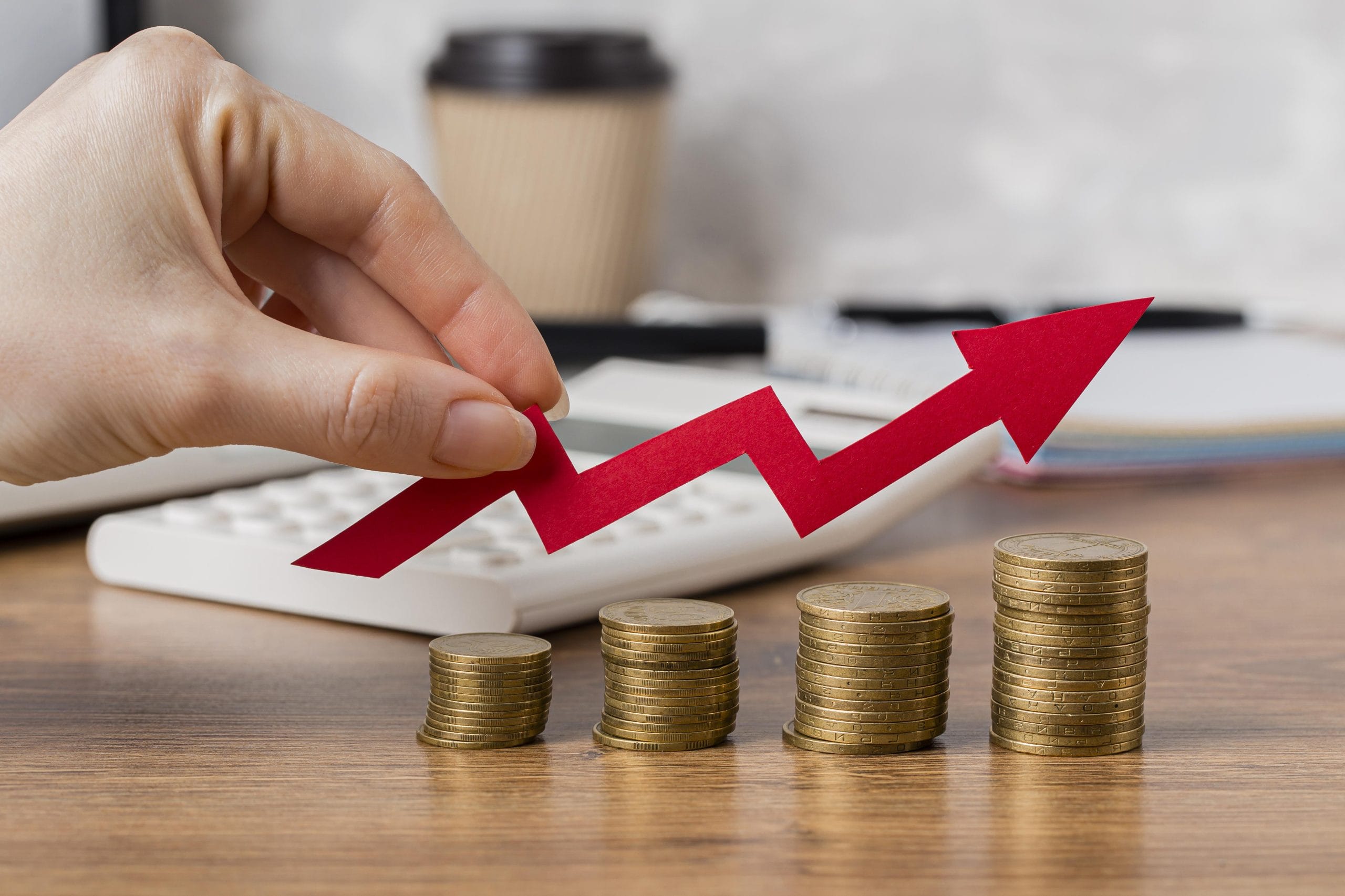 hand-holding-growth-arrow-with-coins A hand holds a red upward arrow above stacked coins, symbolizing financial growth on a desk with a calculator.