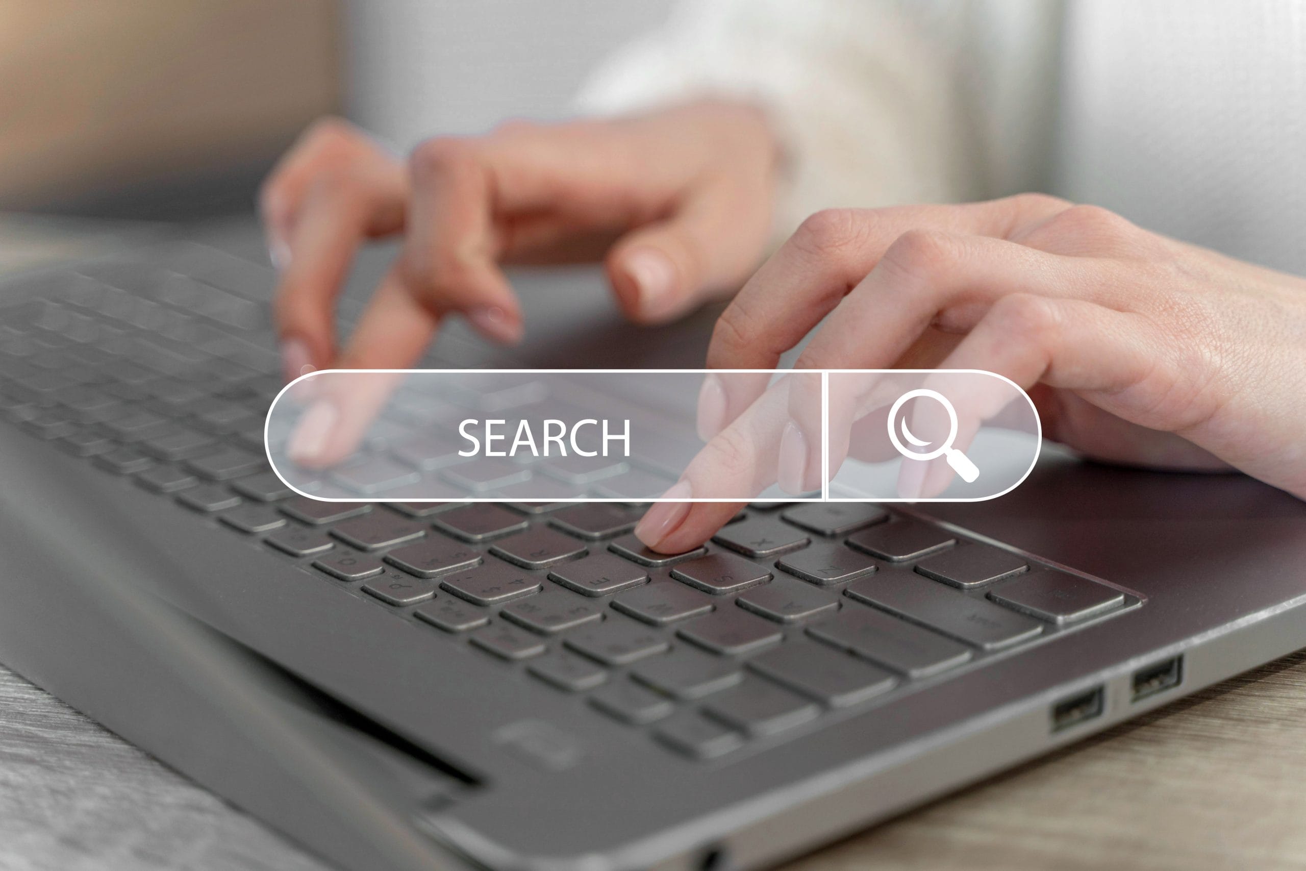 homepage-concept-with-search-bar Hands typing on a laptop keyboard with a transparent search bar overlay displaying the word "SEARCH.