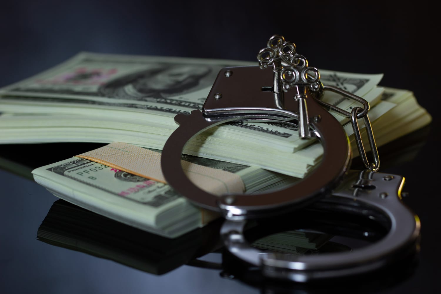 Handcuffs resting on stacks of U.S. hundred-dollar bills on a reflective dark surface.