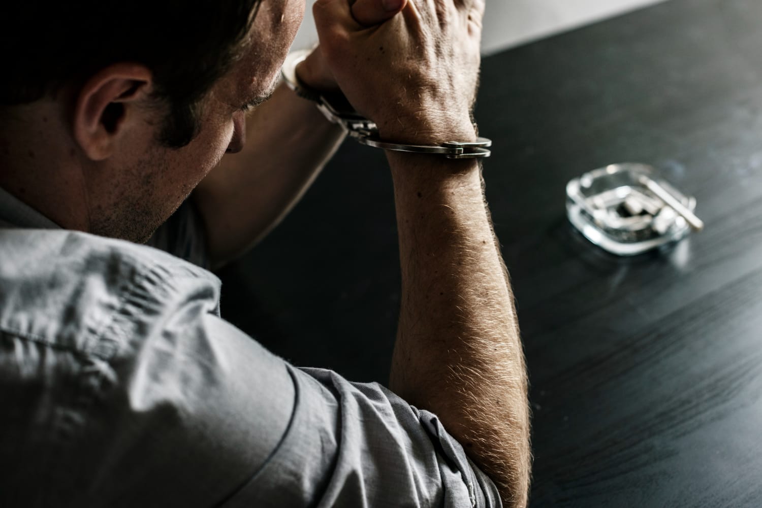 A man in handcuffs sits at a dark table with an ashtray and keys in the background.